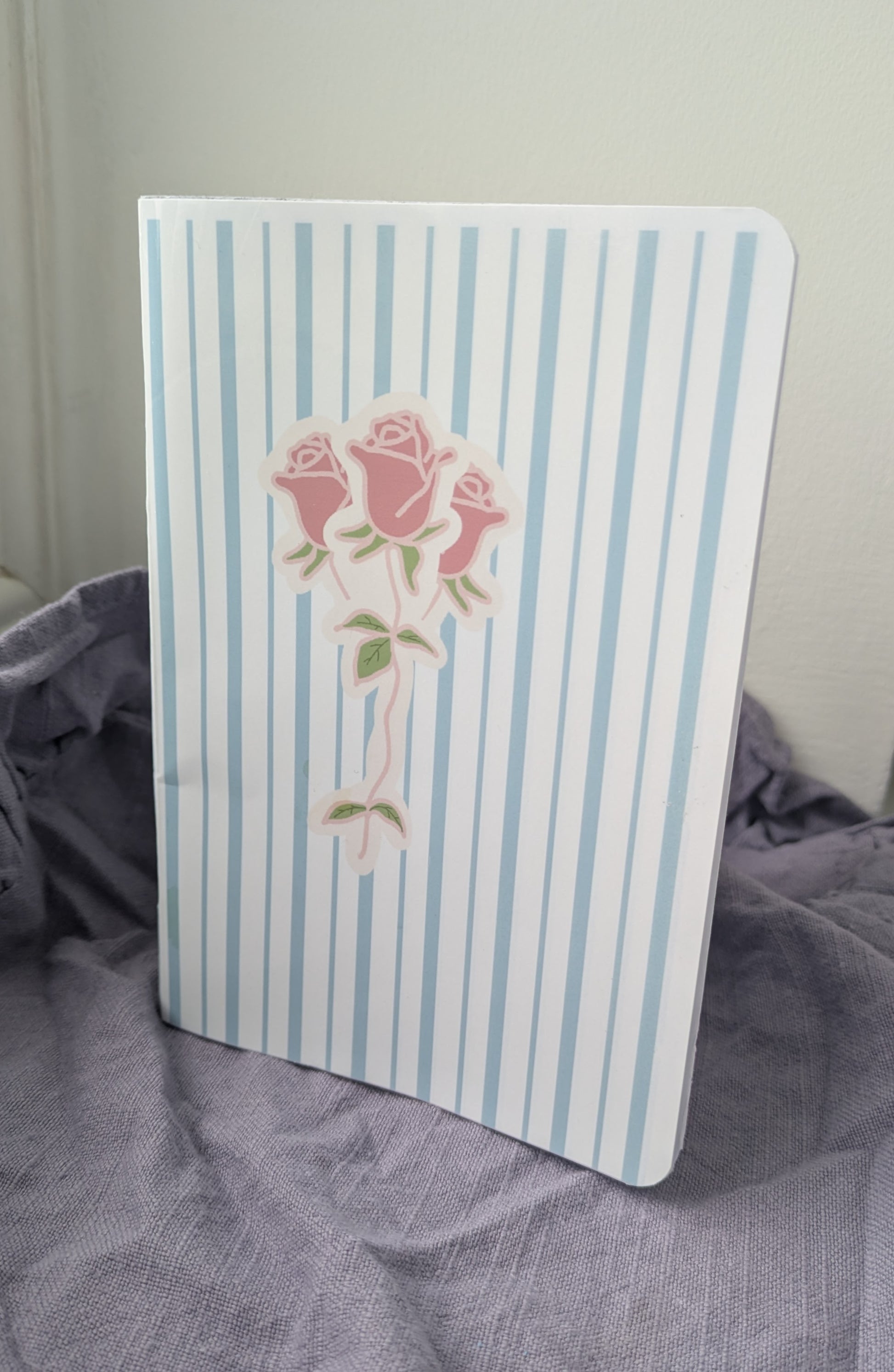 Notebook with blue and white striped cover featuring a pink rose design on a gray fabric background.